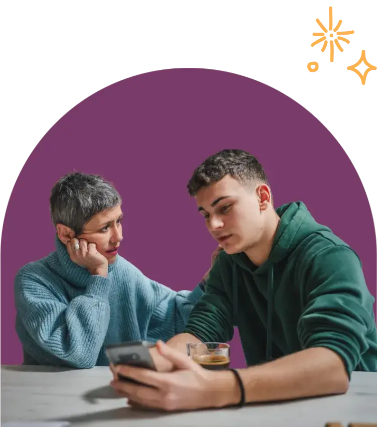A person with short gray hair comforts a young person in a green hoodie, who is holding a phone and learning how to deal with cyberbullying. They sit at a table with a glass on it. The background features purple and abstract yellow shapes.