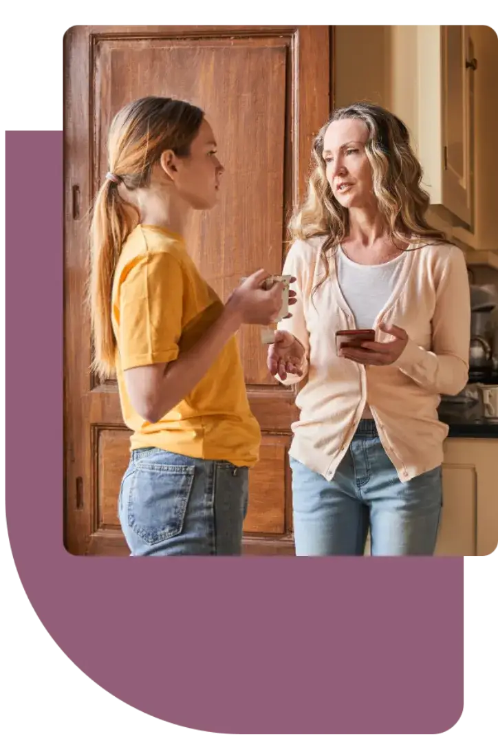 Two women are standing in a kitchen, chatting. One wears a yellow shirt and holds a mug, while the other dons a pale cardigan, clutching her phone. With the Youth Slang Dictionary soundtracking their convo, they engage deeply against the backdrop of a wooden cabinet.