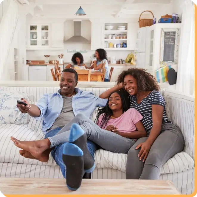 A family of three sits on a couch, smiling and watching TV, mindful of the impact of screen time. In the background, another person is in the kitchen, visible through the open-plan design. The room is bright and cozy, with white walls and modern decor.