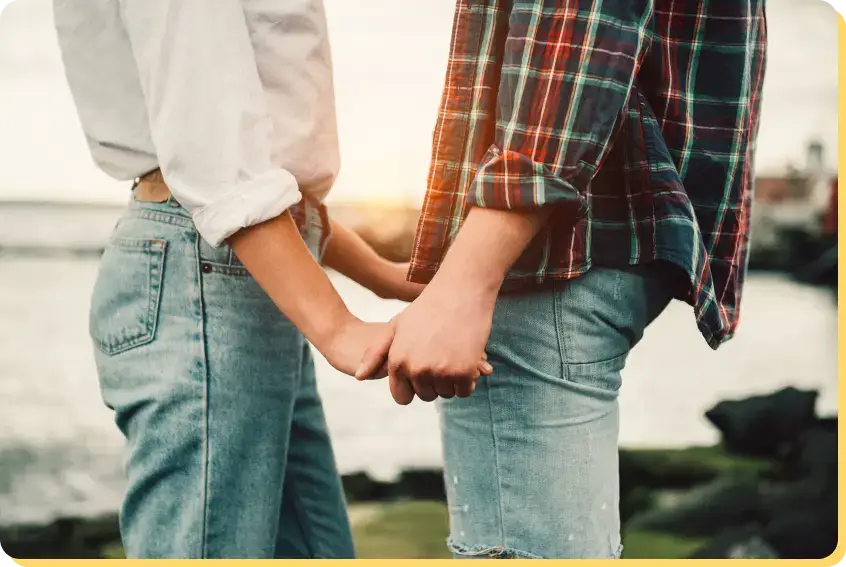 A couple stands face to face by the water, holding hands. The person on the left wears a white shirt and jeans, while the person on the right is in a plaid shirt and jeans. The sunset light creates a warm glow in the background, offering a stark contrast to the isolating effects of porn addiction.