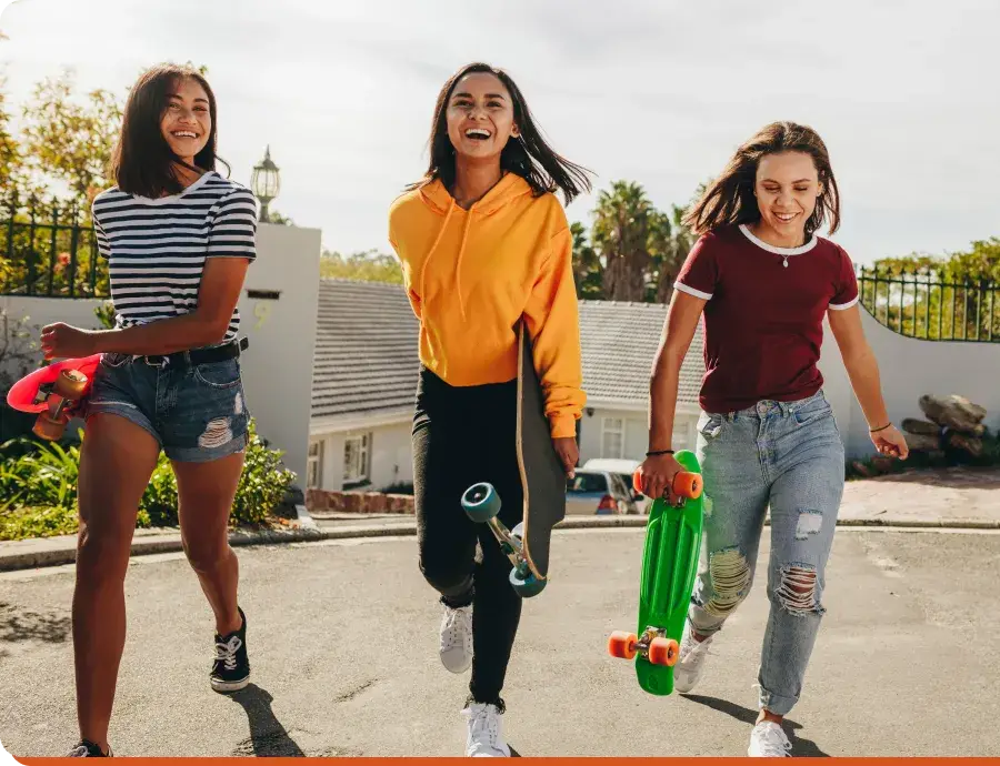 Three friends are cruising down the street, skateboards in hand, sharing laughs and the latest from their Youth Slang Dictionary. One sports a striped shirt, another rocks a yellow hoodie, and the third flaunts a maroon top, soaking up the sun in their vibrant residential spot.