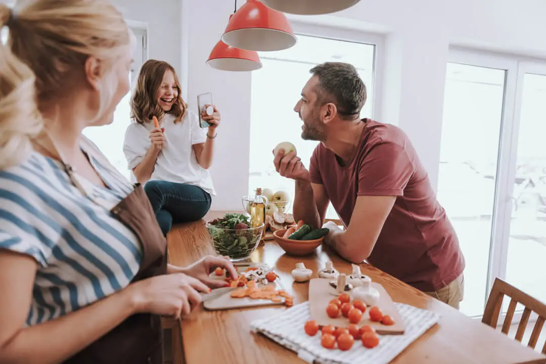 Three people are in a bright kitchen. A woman cuts vegetables on a wooden counter; another sits on it, showing a phone, while a man holds an onion, smiling. Surrounded by fresh veggies and kitchen items, their camaraderie reflects our mission of bringing joy to everyday moments.