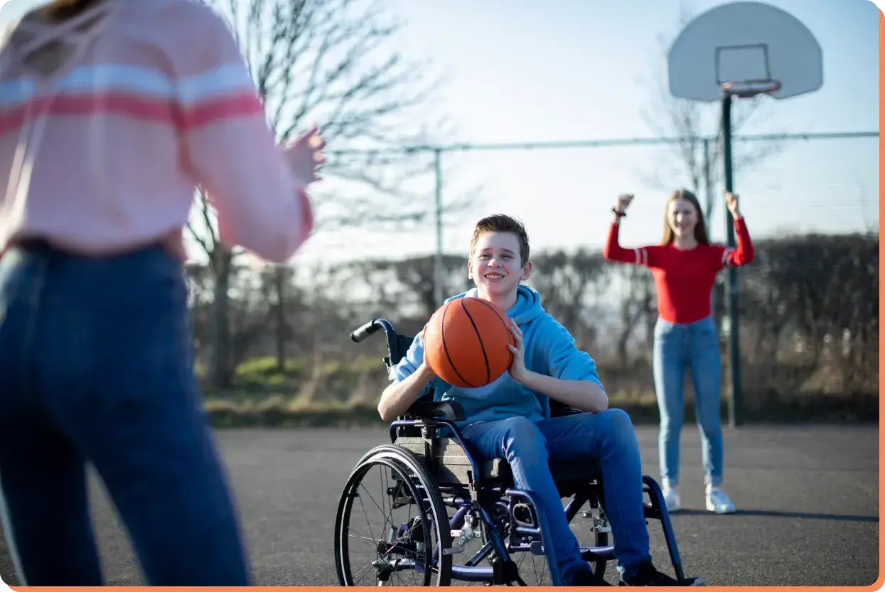 A young person in a wheelchair holds a basketball, smiling and playing with two friends on an outdoor court, far from the impact of screen time. One friend prepares to throw the ball, while the other cheers in the background. A basketball hoop stands nearby.