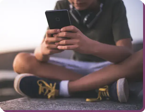 A person sits cross-legged outdoors, holding a smartphone, perhaps exploring topics like the effects of porn addiction. Theyre wearing a green shirt, white shorts, and black sneakers with yellow laces. Black headphones rest around their neck while the background remains blurred.