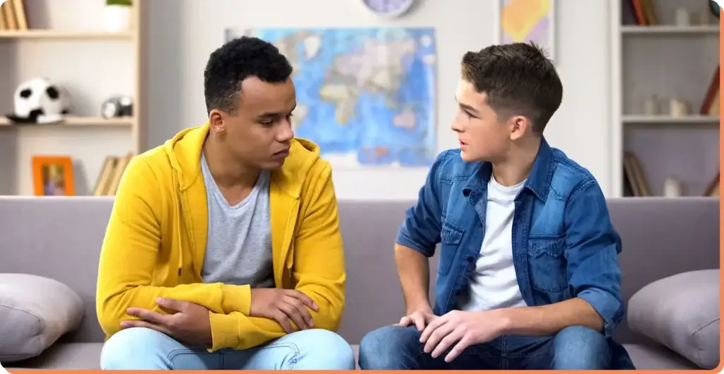 Two young men sit on a couch in a living room, deeply engaged in a serious conversation about the impact of screen time. One wears a yellow hoodie, the other a denim jacket. A map and books are visibly scattered in the background, hinting at their scholarly debate.