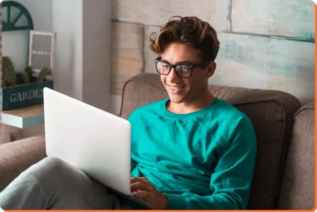 A person with glasses and a teal sweater sits on a couch, smiling while using a laptop to explore teen mental health. A potted plant is visible in the background.