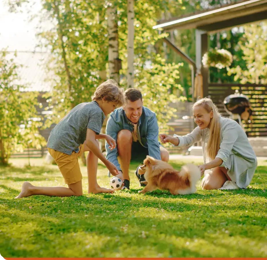 A family of three, a man, woman, and child, play with a small dog in their sunny backyard. The child holds a soccer ball while the adults laugh and crouch. Nearby, a bicycle rests by the wooden fence—an idyllic scene untouched by the impact of social media on teen mental health.
