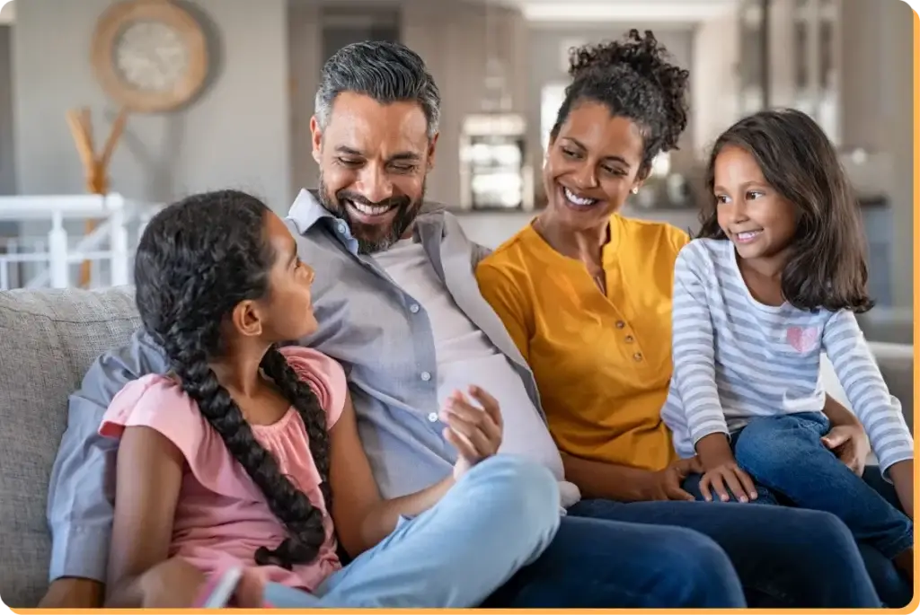 A family of four sits together on a couch, smiling. The father and mother are seated in the middle, with their two young daughters on either side. The cozy living room vibe feels like something from a Youth Slang Dictionary, totally capturing the chill essence of relaxed family time.
