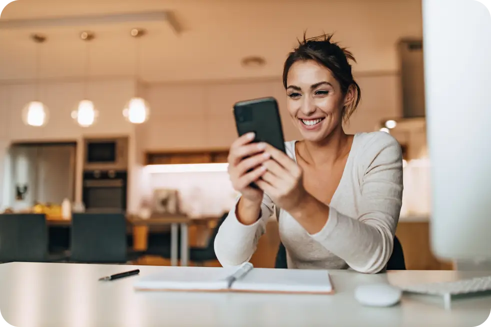 A woman sits at a table in a modern kitchen, smiling at her phone. She wears a light-colored sweater and has a notepad, pen, and laptop in front of her. Warm lighting creates a cozy atmosphere as she prepares to join the movement online.