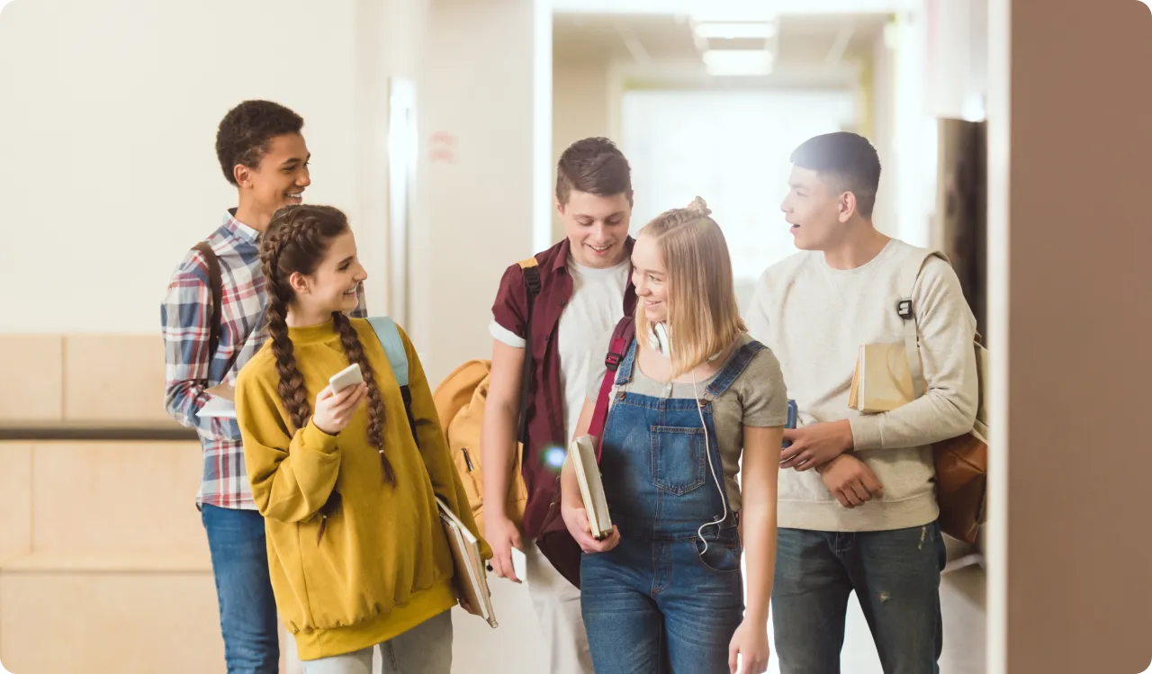 A group of five students strolls down the school hallway, chatting and smiling. Their backpacks swing as they carry books, dressed casually in jeans, a plaid shirt, and overalls. The well-lit corridor is lined with lockers, perhaps hiding their secret youth slang dictionary.