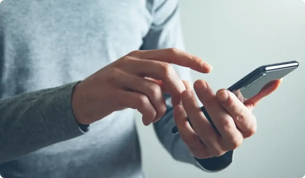 A person in a gray long-sleeve shirt uses a smartphone, holding it with one hand while tapping the screen with the other. The plain light background subtly underscores the hidden struggles many face, such as grappling with the effects of porn addiction in their everyday lives.