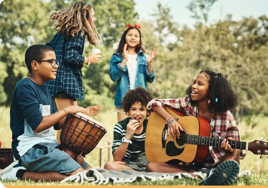 A group of children enjoys music outdoors, free from the impact of screen time. One boy plays a drum, another claps, while a girl strums a guitar. Two girls stand and smile in the background. Theyre sitting on a picnic blanket in a grassy area with trees behind them.