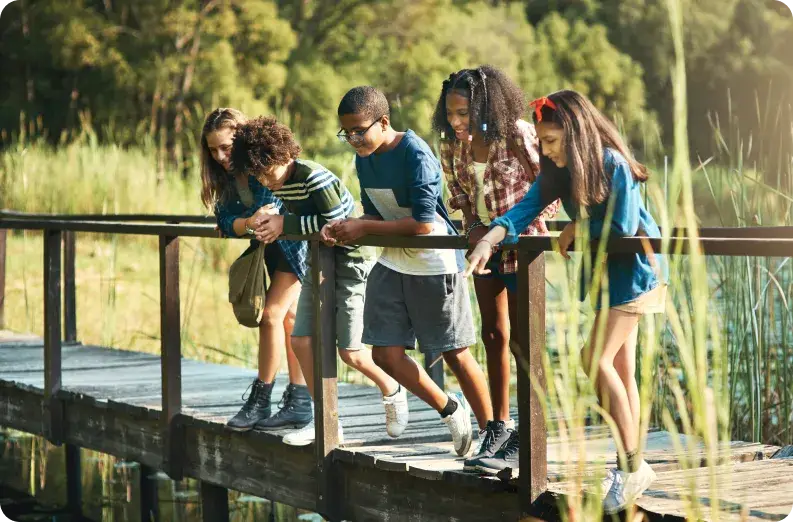 A group of five teenagers stands on a wooden bridge overlooking the water, smiling and leaning over the railing. In this sunny, outdoor setting with lush green foliage, they enjoy each others company away from screens and reflect on the importance of balancing screen time with nature.