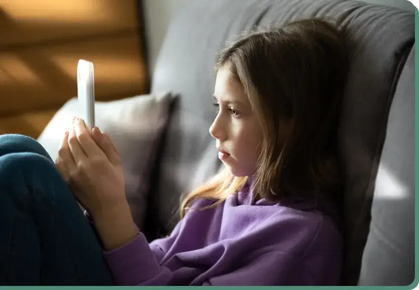 A young girl in a purple sweater sits on a gray couch, intently examining her smartphone, illustrating the impact of screen time. She leans back with bent legs, enveloped in soft lighting, absorbed in the digital glow.