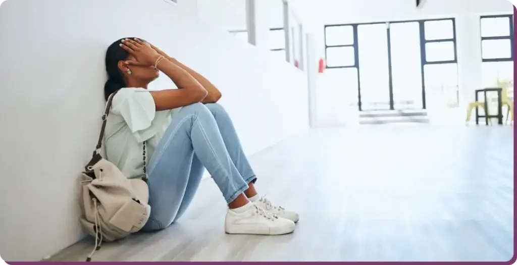 In a brightly lit hallway, a person grapples with the effects of porn addiction, sitting on the floor with hands covering their face. Clad in a light blue shirt, jeans, and white sneakers, they lean against the wall next to a beige backpack.