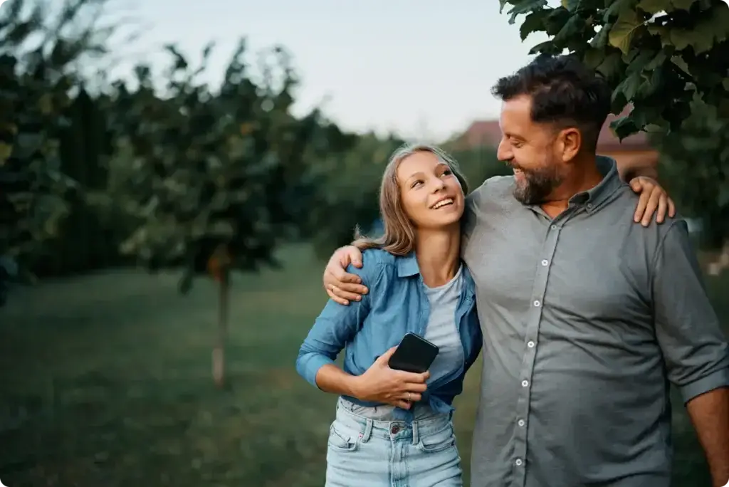 A man and woman stand in a lush garden, smiling warmly at each other. Hes in a gray shirt with his arm around her shoulder, while she holds a phone, clad in a blue shirt and denim shorts. The serene setting contrasts the digital realm, where knowing how to deal with cyberbullying is essential.