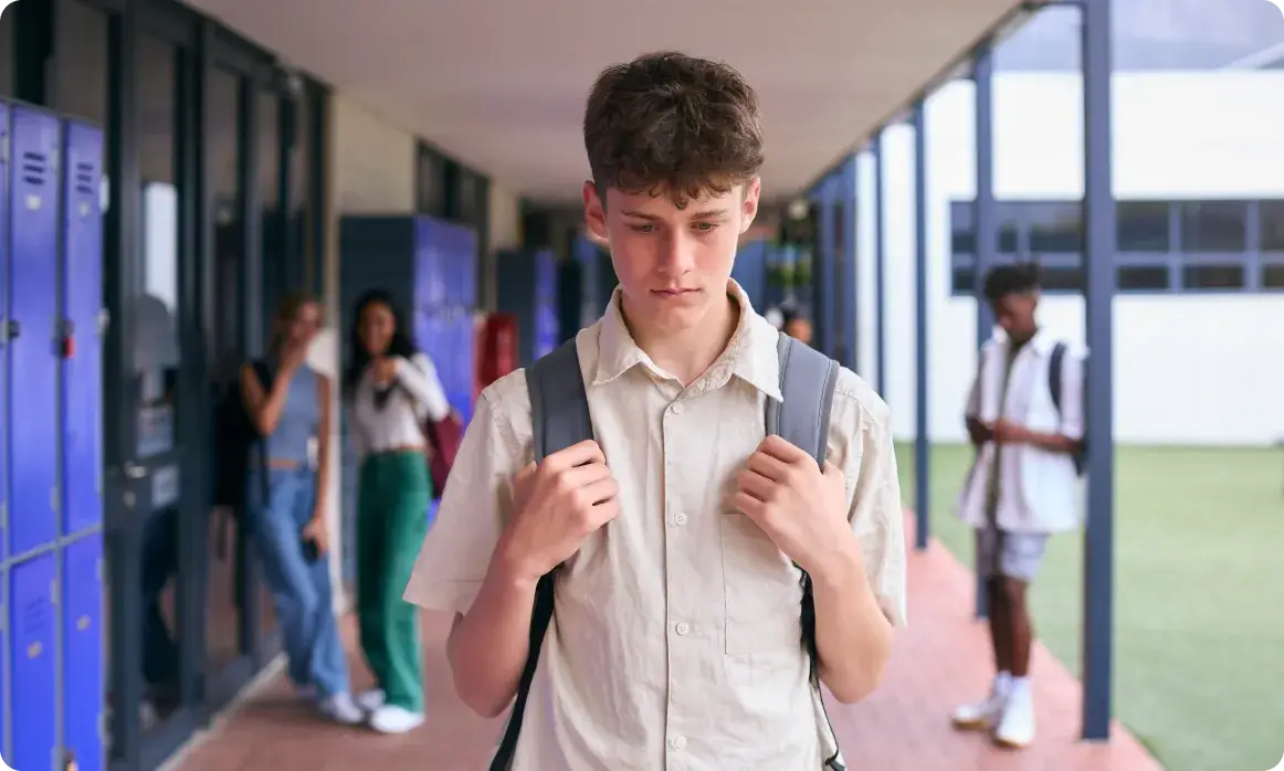 A teenage boy in a beige shirt and backpack stands in a school corridor, contemplating how to deal with cyberbullying. In the background, several other students chat near blue lockers. The setting appears to be a high school.