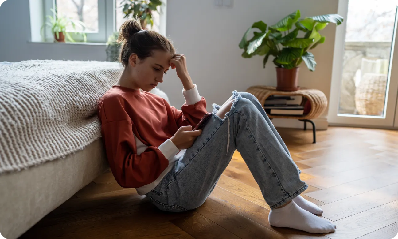A young woman in a red sweatshirt and jeans sits on a hardwood floor, leaning against a bed. She appears focused on a smartphone in her hand. A potted plant and large window are in the background, creating a cozy atmosphere.