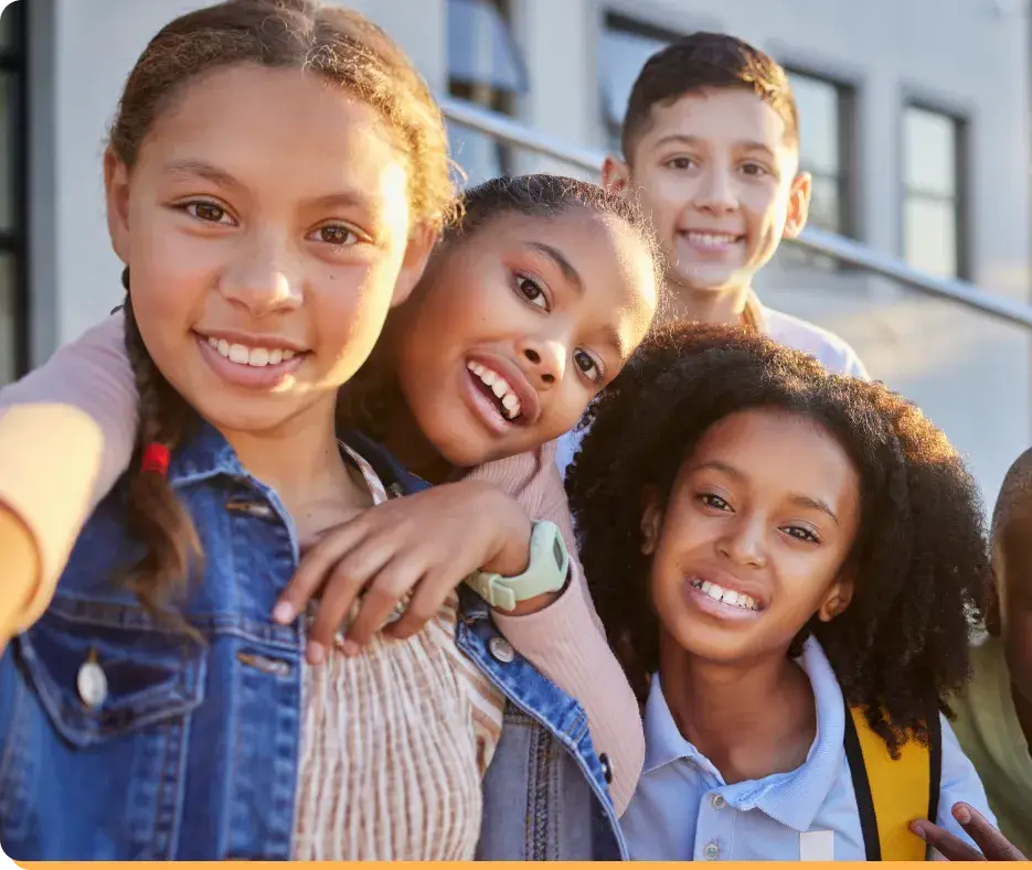 A group of four children, all smiles, pose for a selfie in the great outdoors. Three fill up the foreground while one lingers slightly behind. Against the backdrop of a building bathed in bright daylight, they look like an entry from a living Youth Slang Dictionary. .