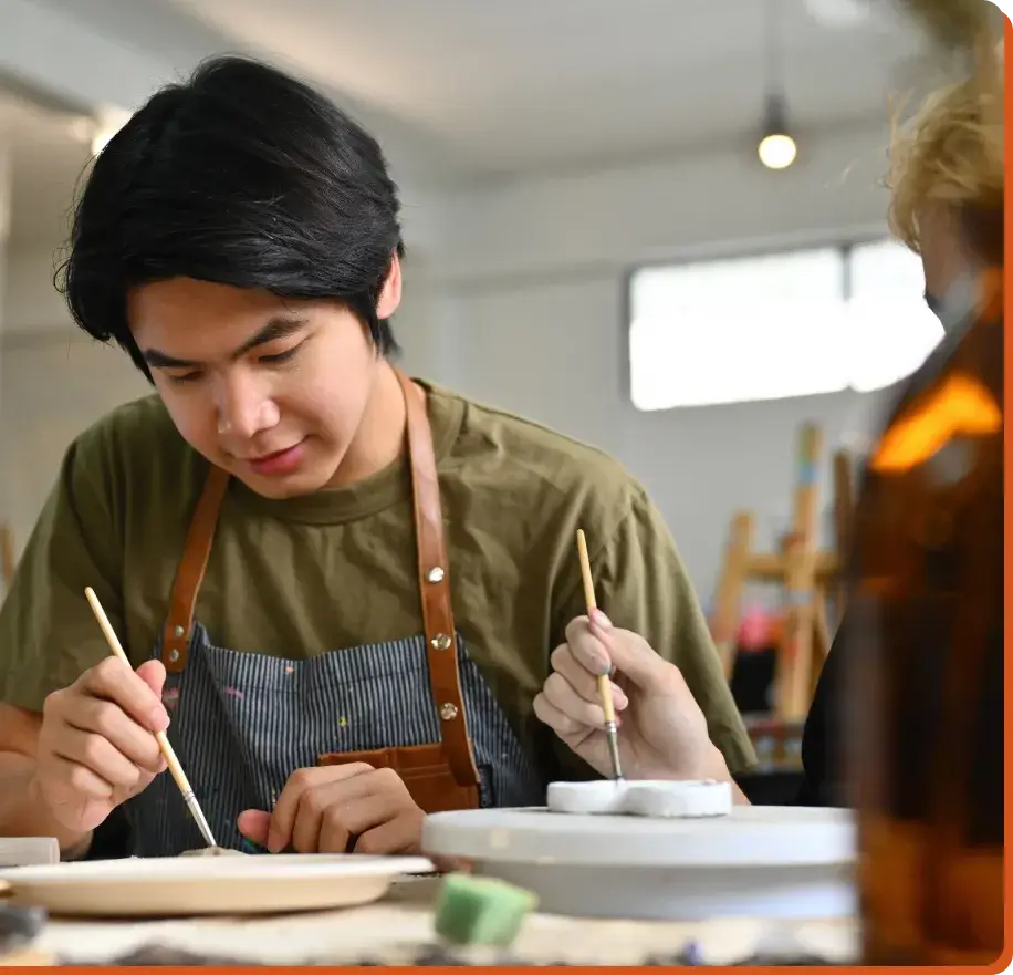 A person wearing an apron is focused on painting ceramics at a table, much like the concentrated focus needed when navigating the impact of social media on teen mental health. They hold a paintbrush, working on a small object while another person is slightly out of focus beside them in the spacious, well-lit studio.
