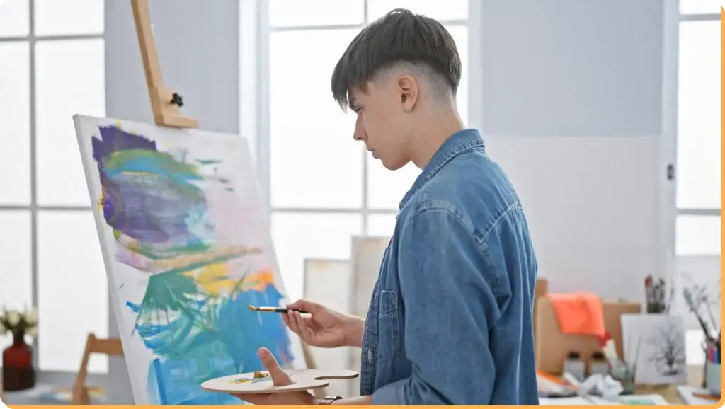 In a sunlit studio, where creativity blooms, a person in a denim shirt paints an abstract piece, vibrant as thoughts on how to deal with cyberbullying. Surrounded by canvases and art supplies, each stroke becomes an expression of resilience amidst the colorful chaos.