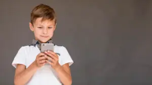 A young boy with short hair is smiling and holding a smartphone in both hands, celebrating Safer Internet Day. He is wearing a white shirt with a dark collar and standing against a plain gray background.