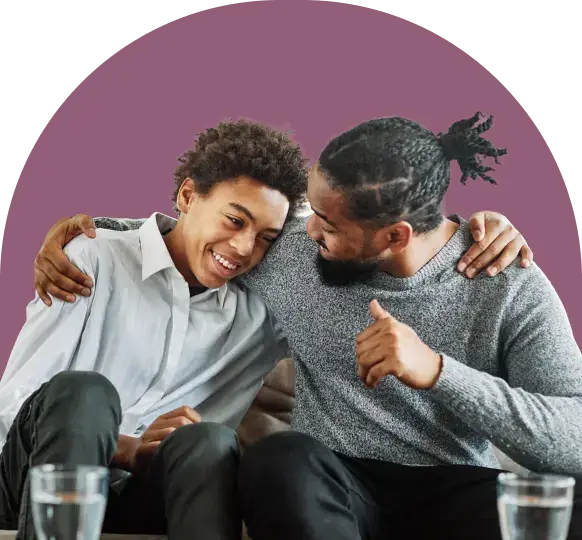 A man and a boy sit closely, sharing smiles and embraces, illustrating a moment of genuine connection that transcends generations. Two glasses of water rest nearby, while the solid mauve background accentuates their bond, subtly hinting at their shared journey in understanding technology together.