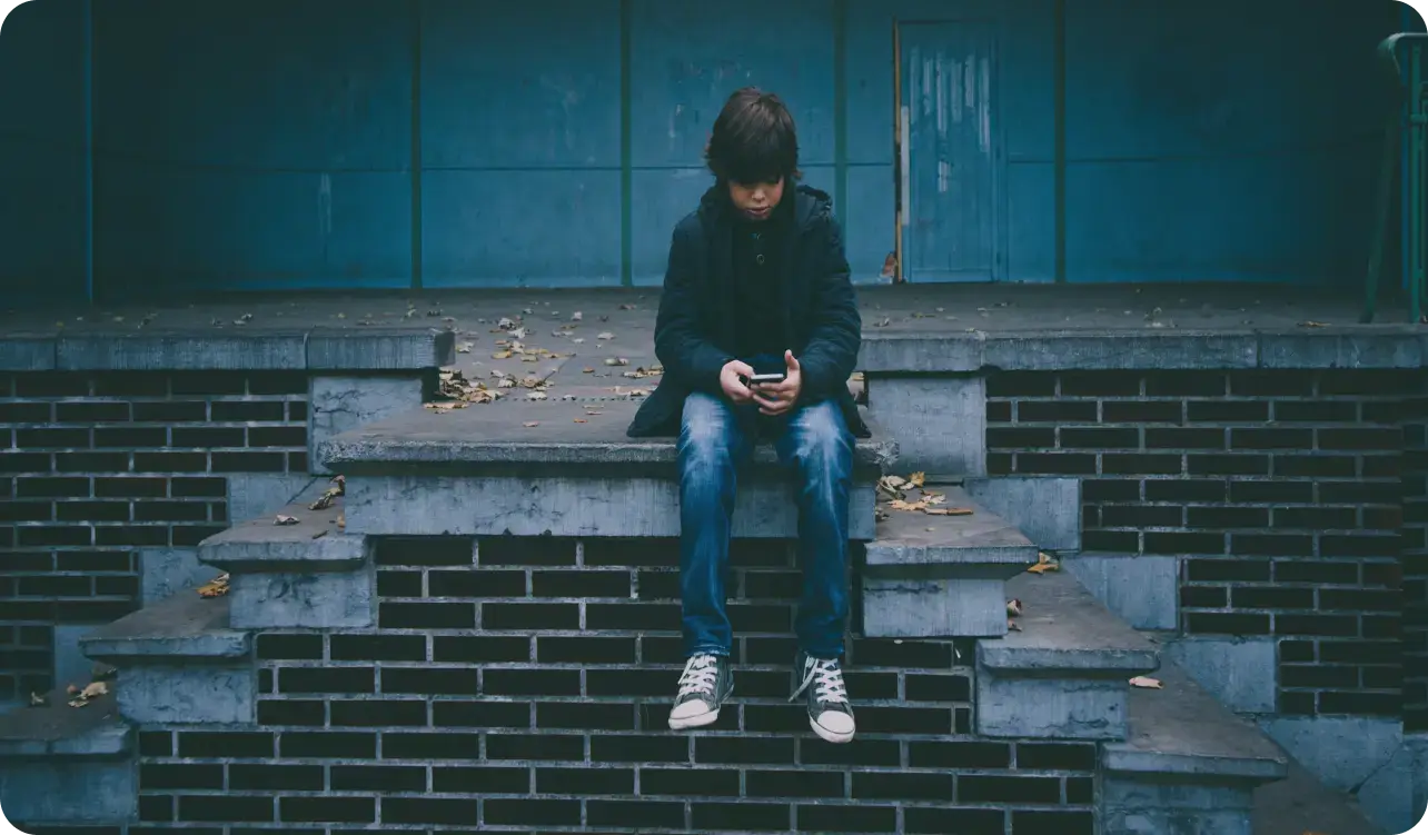 A boy, wearing a dark jacket and jeans, sits on brick steps, absorbed in his phone. Dry leaves scatter around him against a plain, bluish wall. The scene captures a calm moment of introspection as he navigates the world of technology and teen mental health.