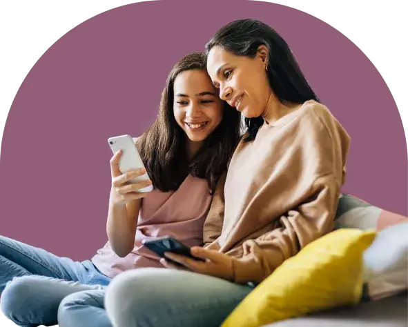 A woman and a young girl sit together, smiling and scrolling through a Youth Slang Dictionary on a smartphone. The woman has long dark hair and wears a tan sweatshirt, while the girl, in a pink top, giggles as they explore new terms on the couch with a yellow pillow.