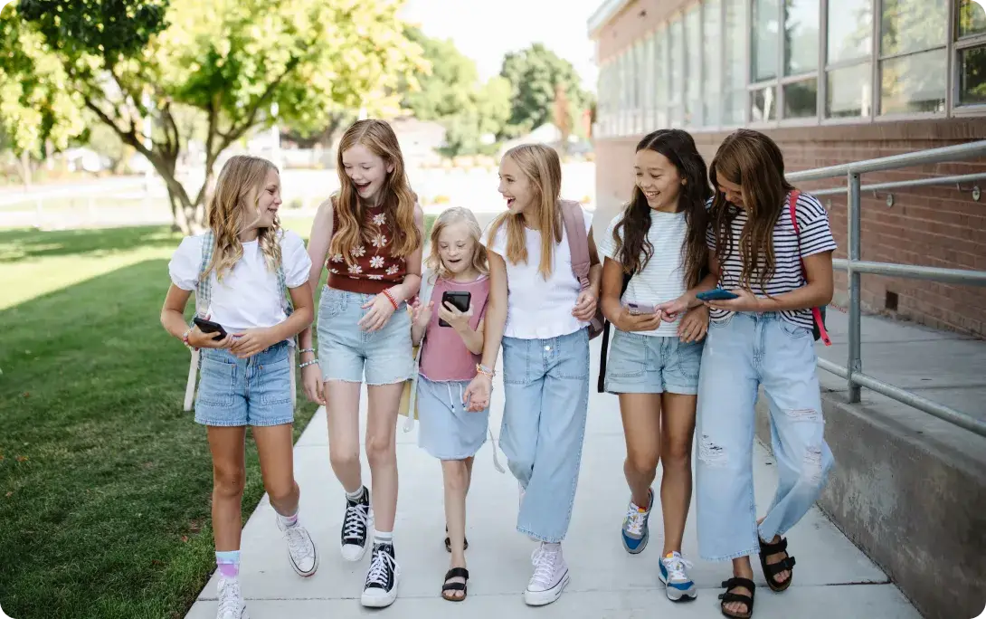 A group of six girls strolls outside, smiling as they check their phones. Casually dressed, they enjoy the sunny day with trees in the background and a brick building nearby. Its a snapshot of friendship in the modern age, subtly highlighting the impact of screen time on social moments.