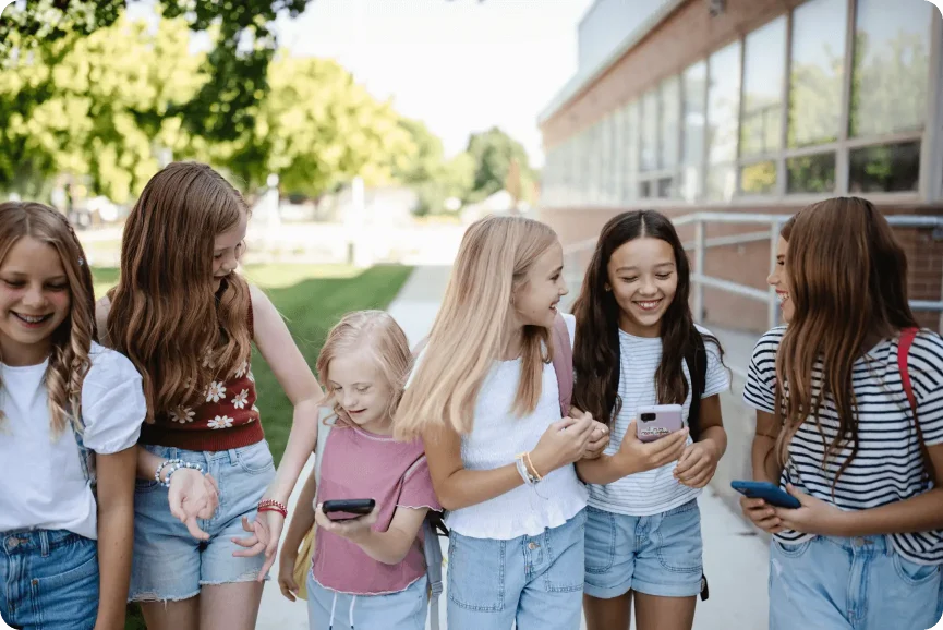 IMG_4710 1 A group of six young girls stand on a sidewalk outside a building, laughing and looking at their Troomi smartphones. They are dressed in casual summer clothes, and the background features trees alongside a glass-windowed building.
