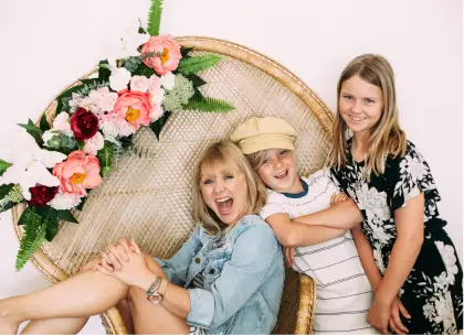 A joyful woman sits in a wicker chair adorned with pink and white flowers, alongside a smiling young child wearing a cap and a girl in a patterned dress. They all share a happy moment in front of a white background.