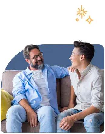 A man with glasses and a beard, wearing a blue shirt, sits on a sofa smiling at a younger man in a light shirt and ripped jeans. They both look happy and relaxed, as if discussing How to Deal With Cyberbullying, with abstract star-like graphics above them.