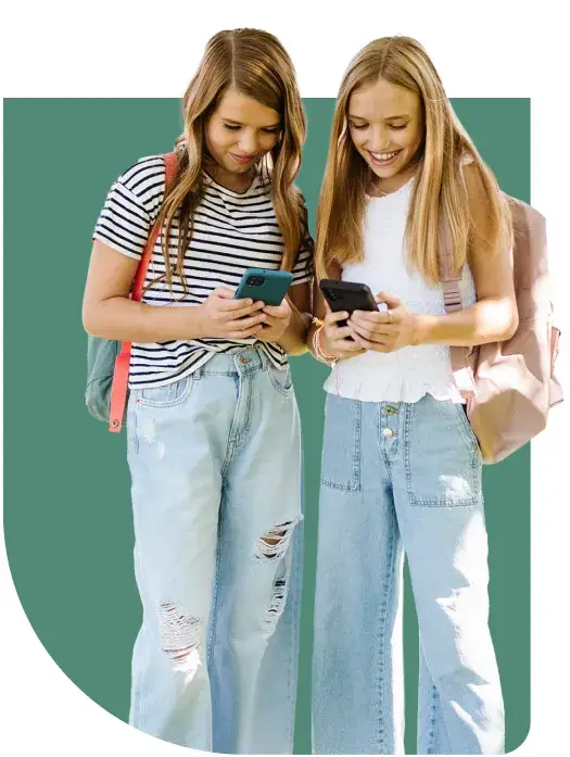 Two young girls standing closely, both holding smartphones and smiling, embody the impact of social media on teen mental health. Theyre wearing casual outfits with denim jeans and backpacks against a solid green background with rounded corners.