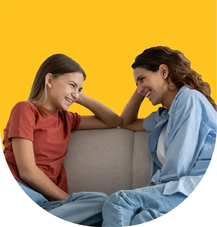 A woman and a girl sit on a couch, smiling and facing each other against a yellow background, embodying the spirit of understanding. The woman has curly hair and wears a blue shirt, while the girl, donning a red shirt, represents the vibrant curiosity of teen mental health.